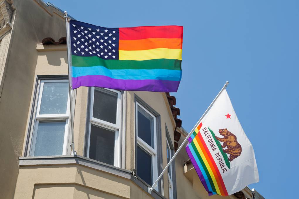 Two flags hang from the bay window of a home. One flag is the California state flag with the rainbow flag on the lower half and the other flag is the US flag with the stripes replaced with a rainbow.