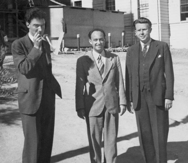 Black and white image of three men in suits standing on the grounds of a college campus.