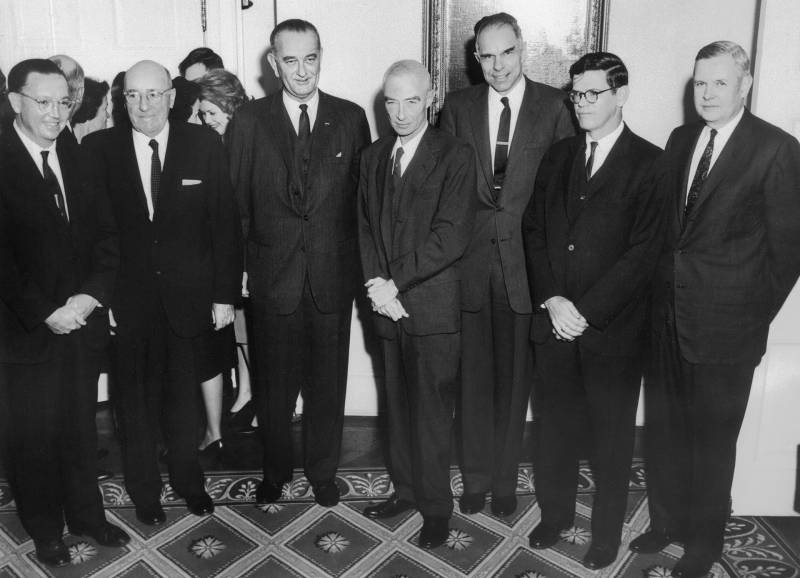 Archival black and white photo of 6 men in dark suits standing in a row inside the White House.