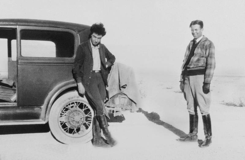 Black and white image. Two men stand in the open desert, with bright sun blazing. One is leaning against the back corner of a 1930s car while the other stand a little off to the right. They are both wearing knee high black leather boots.