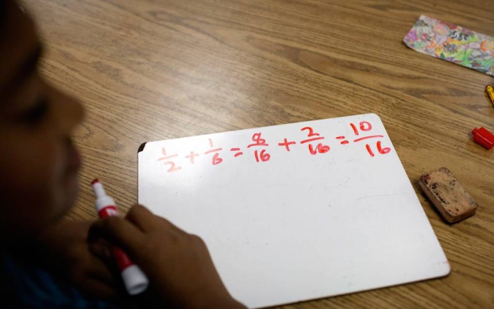 Numbers written in red marker on a small white board. A student is in the foreground holding the red marker.