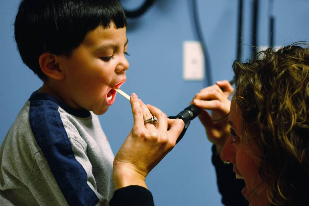 A little boy sits in a health care clinic room where a nurse is looking inside his mouth using a wooden depressor and a light.