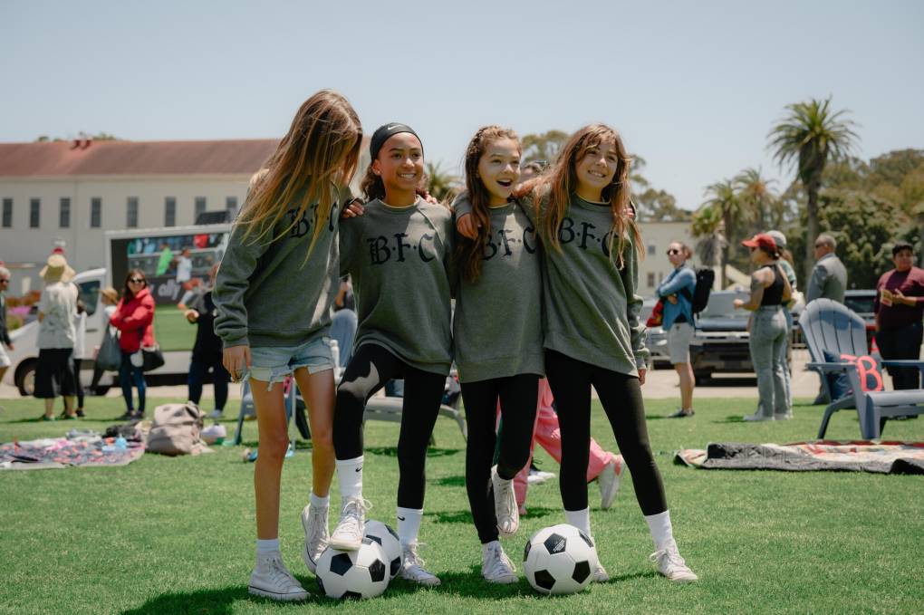 Four your girls in gray sweatshirts stand in a row on a soccer field. Two of them are holding soccer balls under their feet.