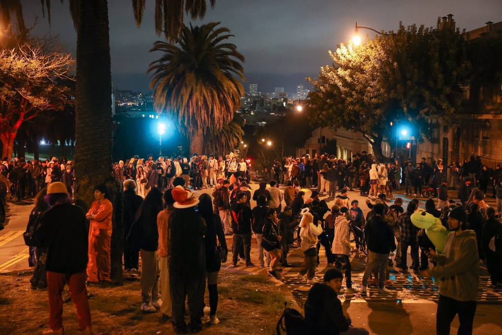 A nighttime image of a palm tree-lined street, with many people gathered under the street lights.