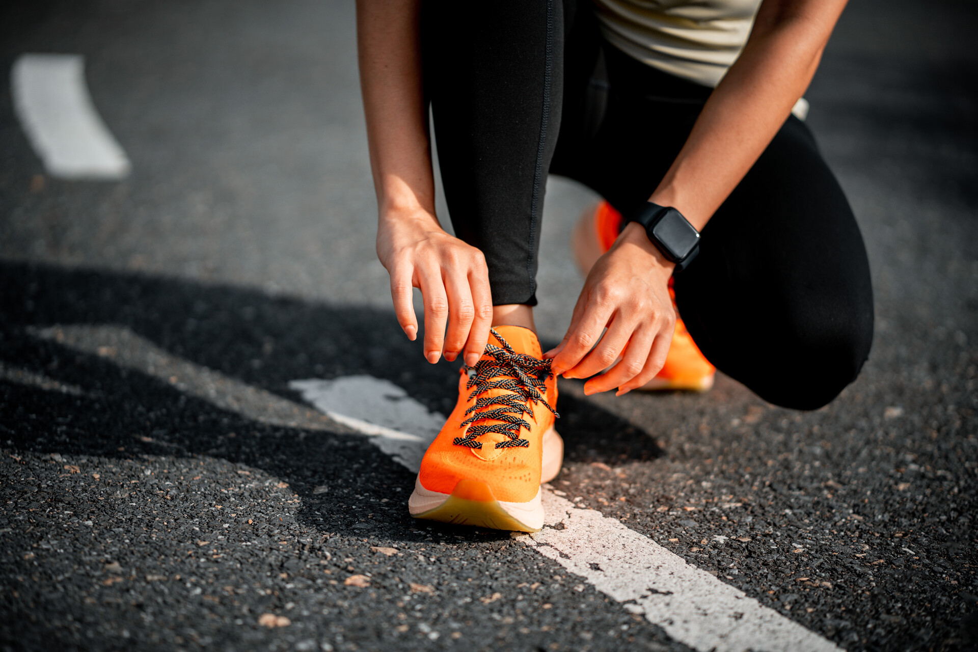 An image of a person with lighter skin crouching with one knee bent to tie the laces of their neon orange running shoes. We can't see their face.