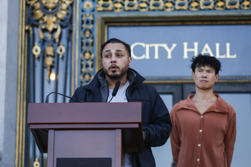 A man wearing a dark hooded sweatshirt stand at a podium with another man standing to his right wearing a reddish shirt in front of a building that has a sign reading "City Hall."