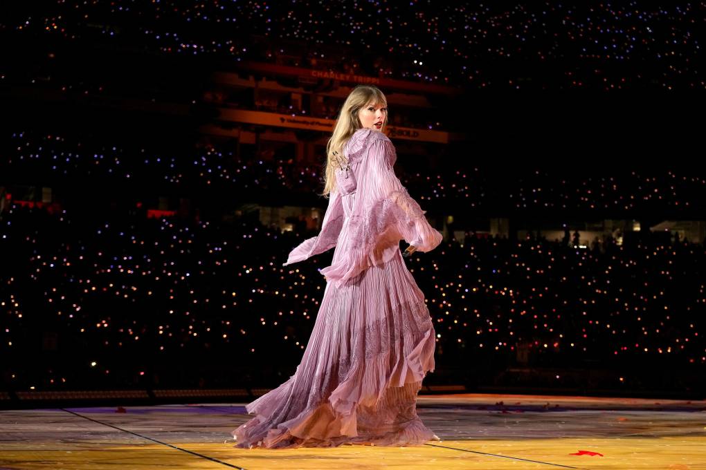 Taylor Swift, a white woman with blond hair, stands on a stage in a pink flowing long-sleeved dress looking backward at the camera.