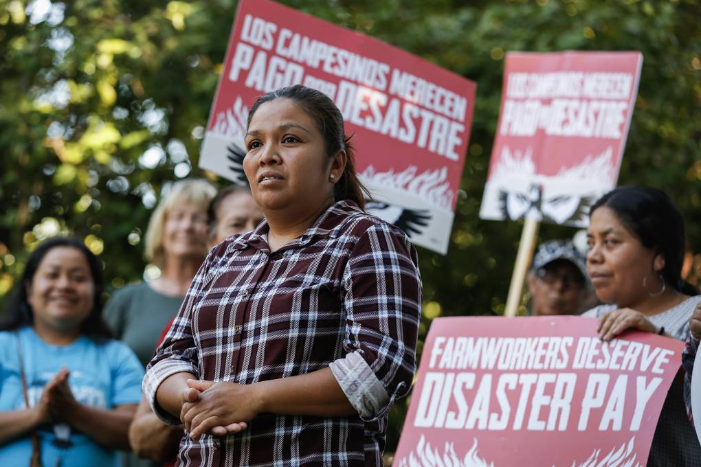 A person with long hair and a plaid shirt stands in front of others holding signs reading "Farmworkers Deserve Disaster Pay" in an outdoor setting.