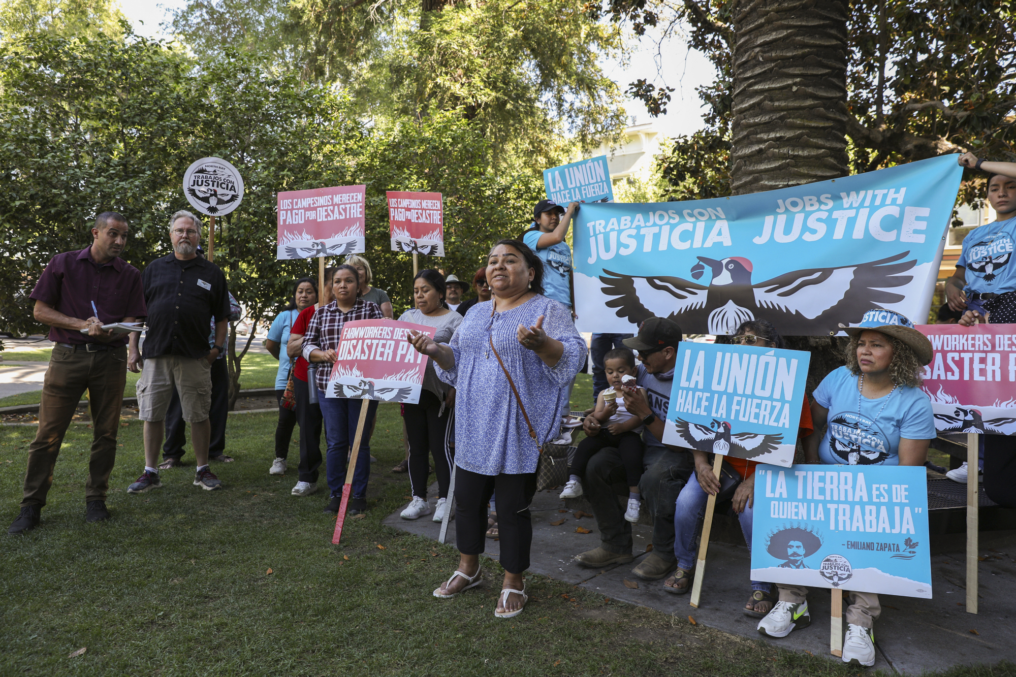 A person wearing earrings speaks in front of others holding signs reading "La Unión Hace la Fuerza" and "Farmworkers Deserve Disaster Pay" in an outdoor setting.