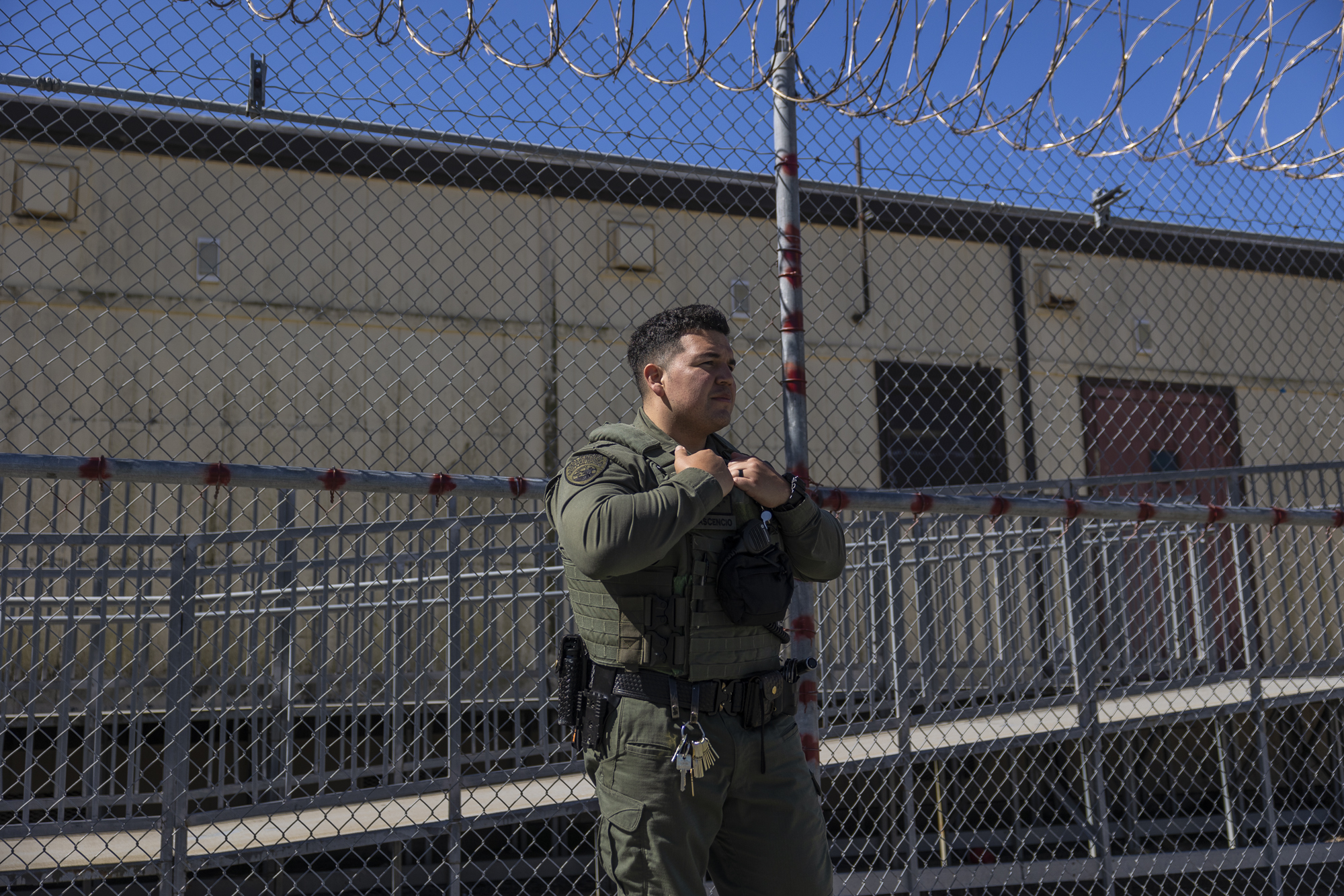 A person in a green uniform stands in front of a chain link fence topped in barbed wire.