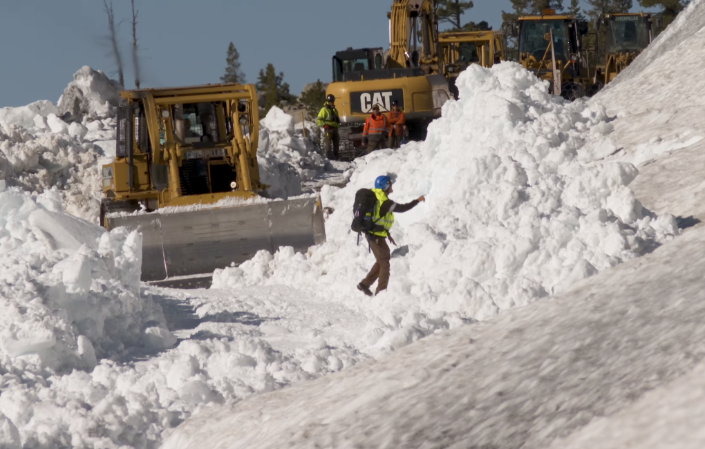 A worker in hardhat and backpack directs bulldozer operators working to clear piles of snow from mountain road.