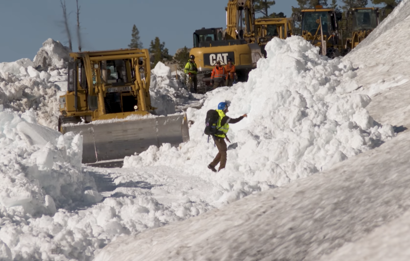 A worker in hardhat and backpack directs bulldozer operators working to clear piles of snow from mountain road.