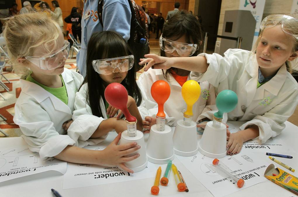 A group of four elementary school-aged girls are wearing white lab coats, clear goggles as they participate in a science experiment with styrofoam cups, balloons and air. A box of Crayola crayons are scattered on the table. The little girls' lab papers are also on the table.