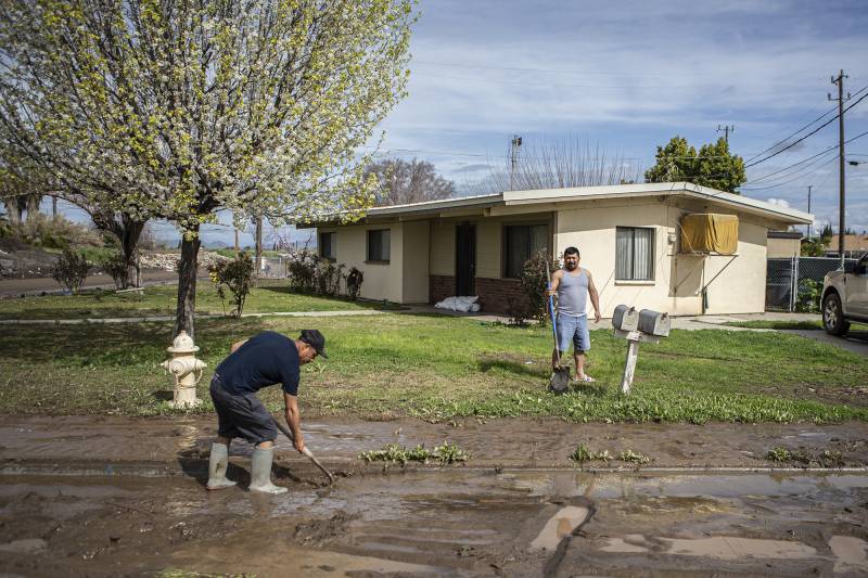 Two men are seen in the front yard of a house. The man to left is wearing a black tshirt, cap, and rain boots shoveling mud while the man on the right in a grey undershirt holds a shovel.