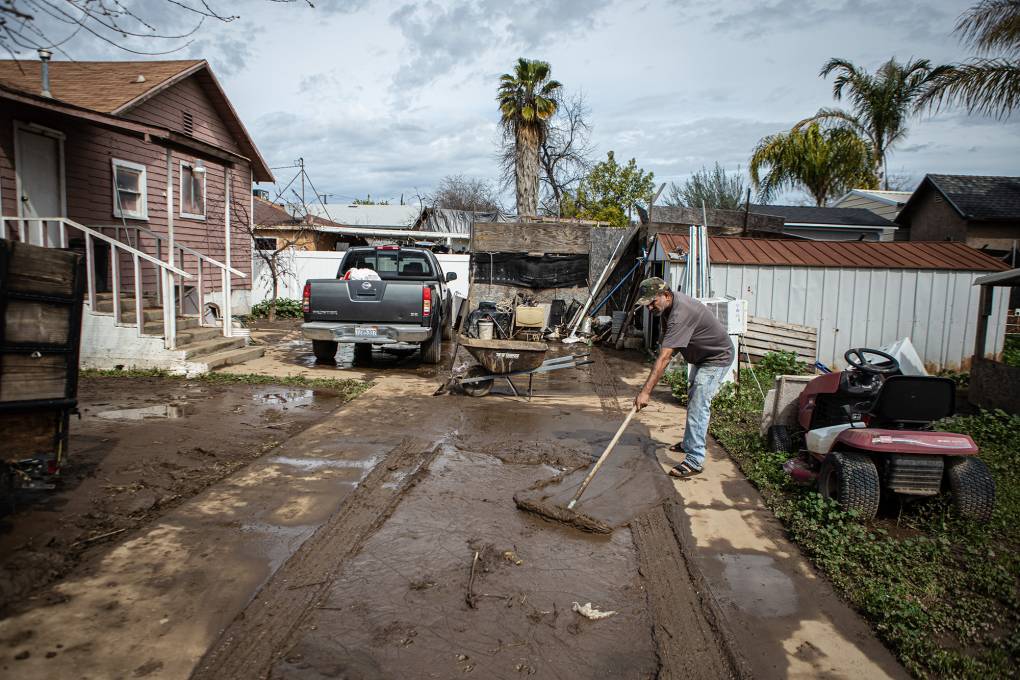 A man wearing a brown t-shirt and a cap pushes mud with a broom in a driveway that has two vehicles, equipment and a house in the background.