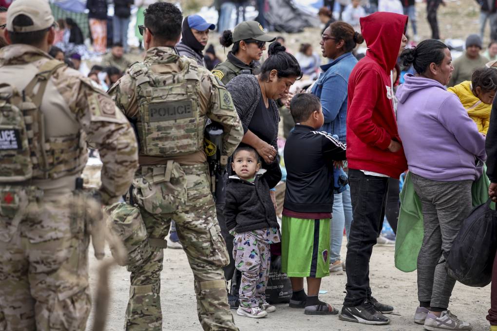 A group of people including children stand in line as people wearing camouflage clothing observe them.