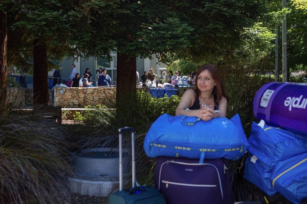 A woman sits with a blue bag on top of a purple suitcase in front of a tree.