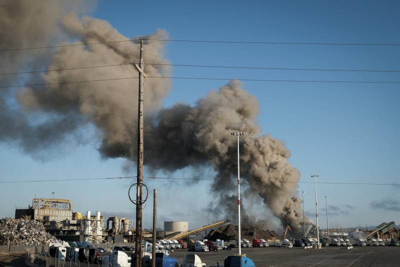 Large clouds of smoke rise from an industrial-looking space where many truck cabs are parked.