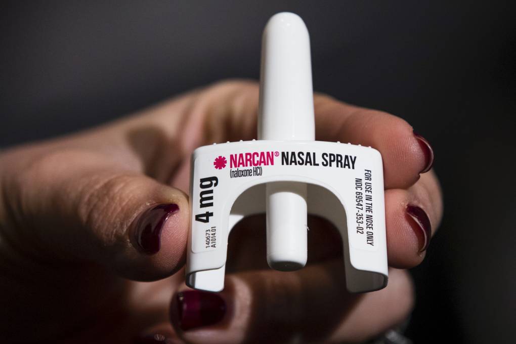 A hand with painted finger nails holds a small white object with the words "NARCAN NASAL SPRAY" written on it.