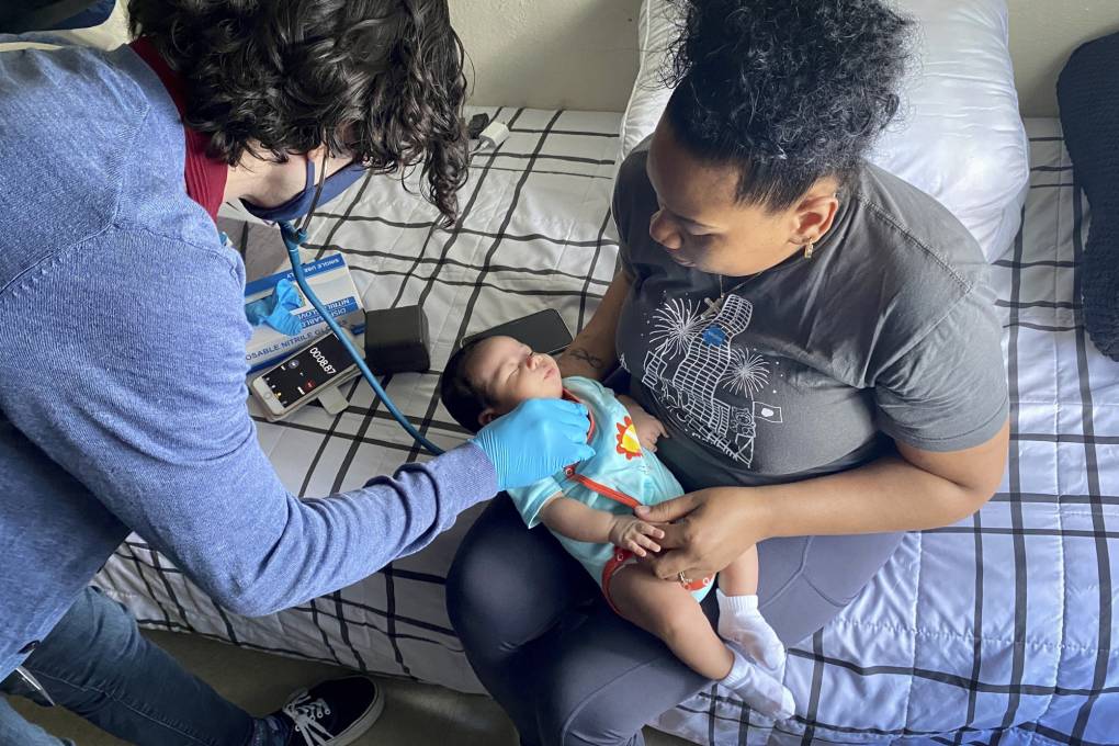 A person cradles a baby in their lap while sitting on a bed as a person wearing medical gloves uses a stethoscope on the baby.