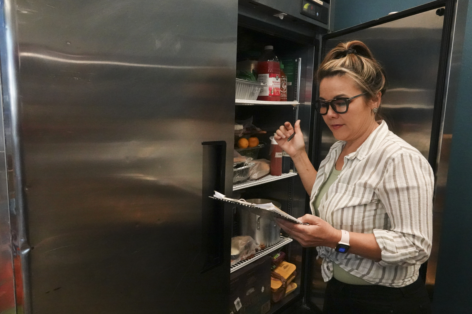 A woman with light-brown hair and black glasses holds a clipboard in one hand, a pen in the other, as she stands in front of a large refrigerator filled with food.