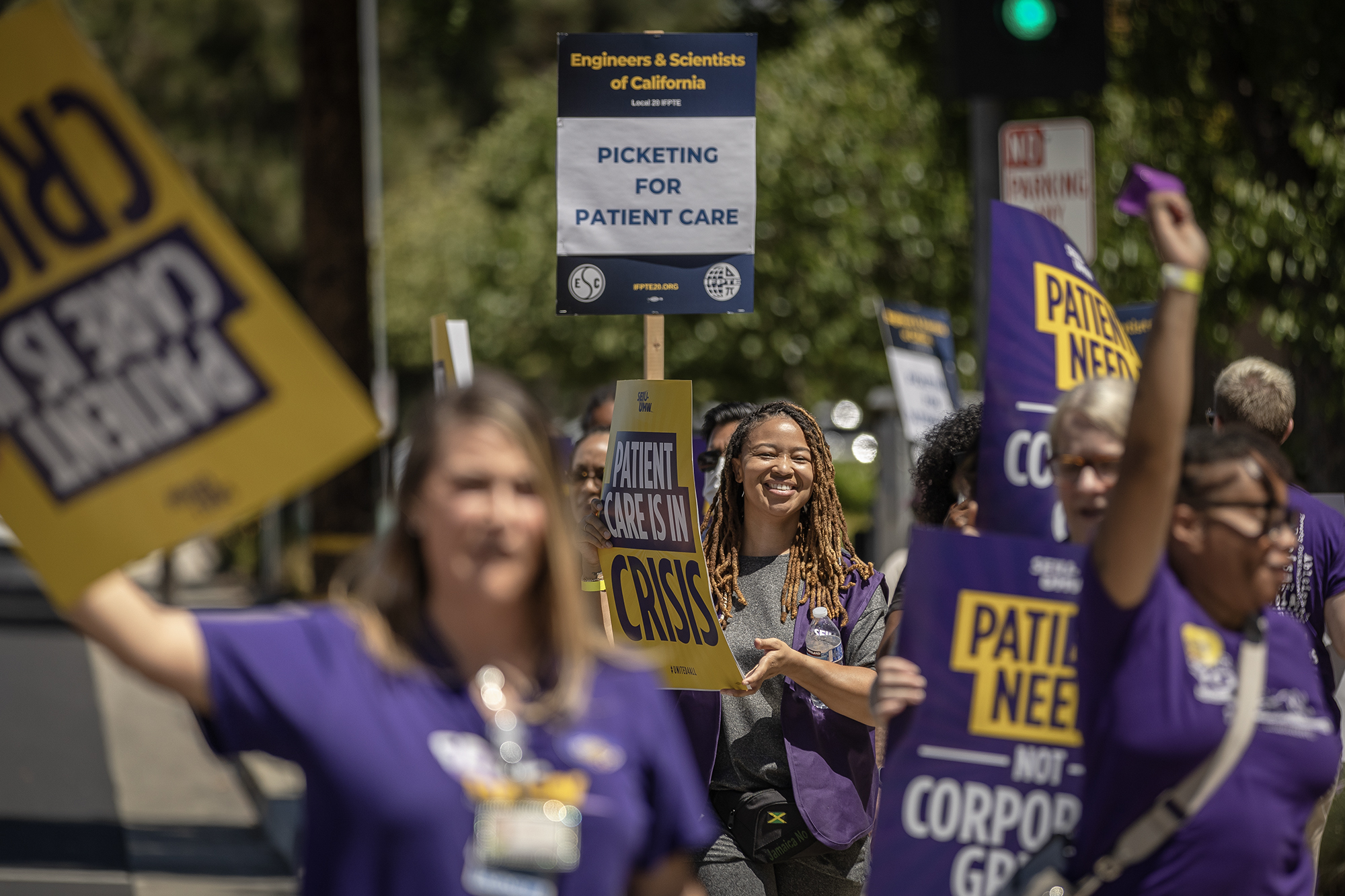 Dozens of health care workers march in protest outside of a Kaiser Permanente building in Sacramento, California. One yellow and black sign reads, "Patient Care Is In Crisis."