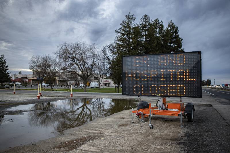 An electric road sign on a gloomy, wet day reads, "ER and Hospital Closed." A hospital is seen in the background with several white vehicles in the parking lot.