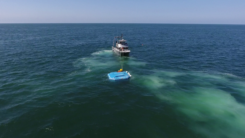 A boat sits on the open ocean, tugging a large, blue, metal rectangular machine into place.