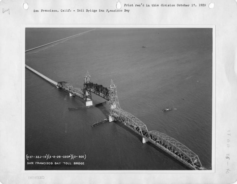 A black and white archival aerial photo of the middle section of a long bridge passing over the San Francisco Bay.
