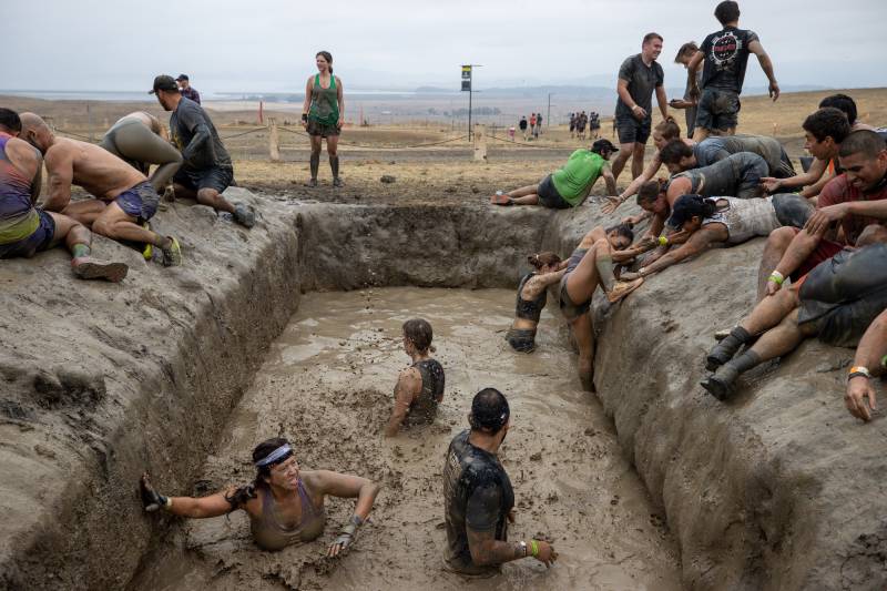 a dozen people, dressed in athletic gear, climb in and out of a muddy pit in a field