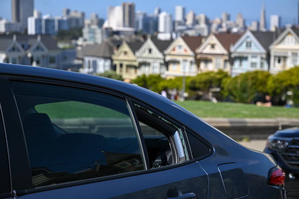 A car is parked next to a large park with a view of the San Francisco skyline. The rear window on the left side of the car has been shattered.