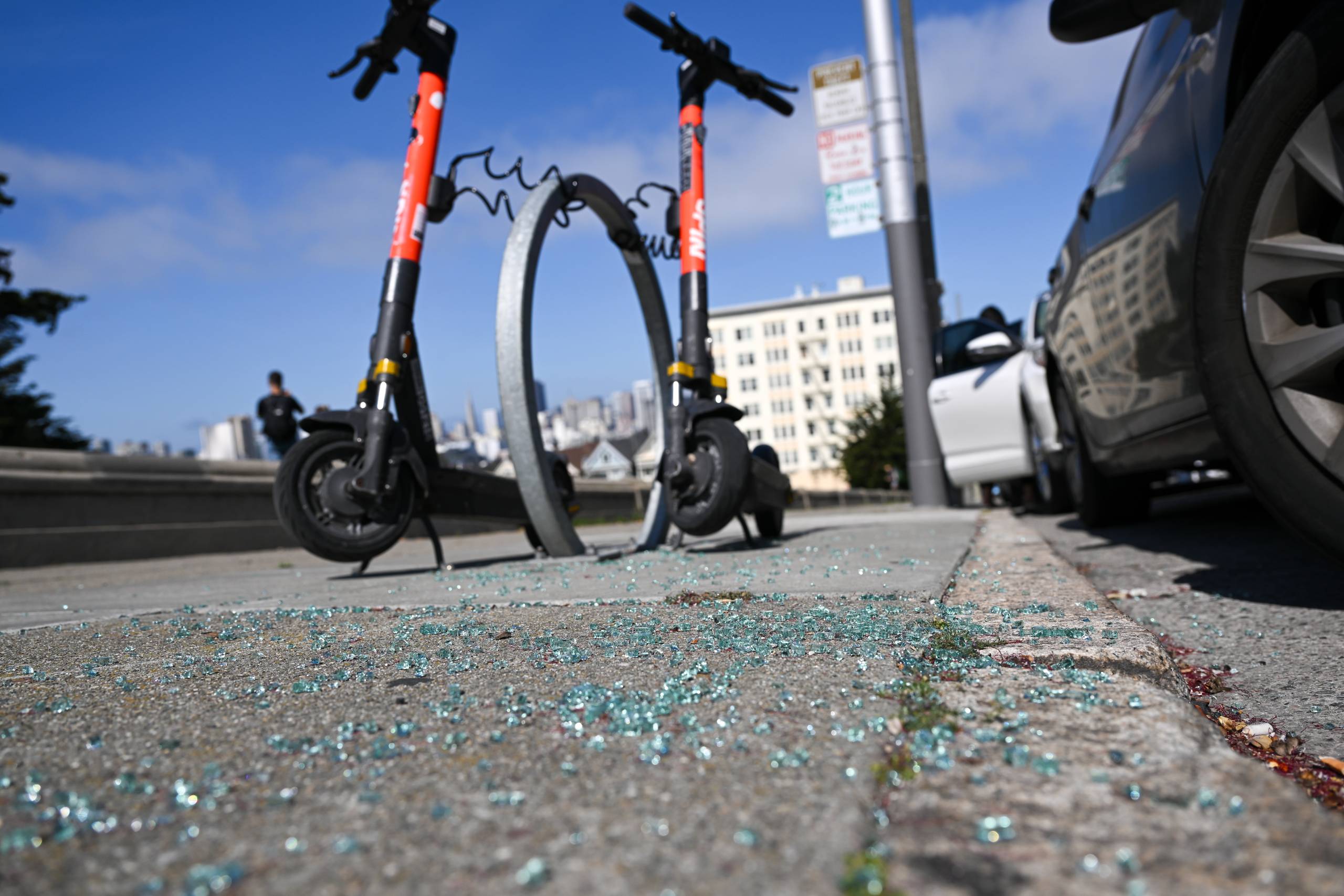 Shattered glass from a car window covers a street sidewalk. There are two electric scooters parked nearby.