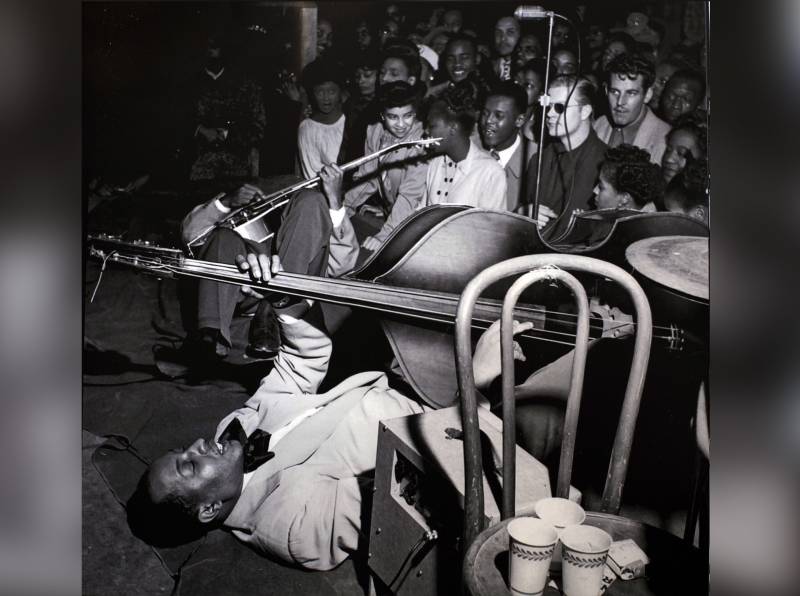 Black and white image of a man in a light color tuxedo playing upright bass while laying down on a stage. A crowd of people stand listening just in front of the stage. A chair with cups sitting on it is visible in the foreground.