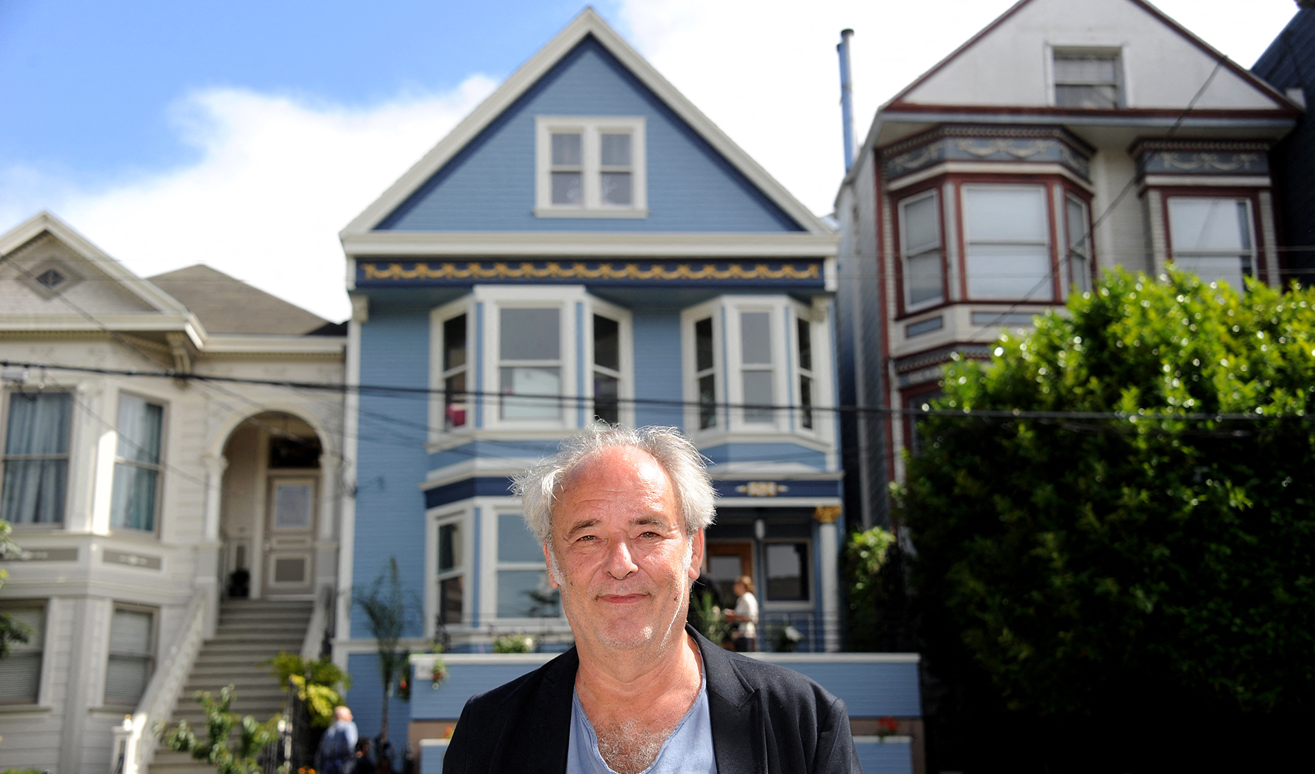A man stands centered in front of a blue Victorian home. He is framed by the house. He has white hair and is wearing a blue shirt with a black blazer over top. 