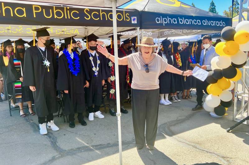 A woman wearing a hat with her arms outstretched stand behind students in a graduation gowns under a tent that reads "Public Schools Alpha Blanca Alvarado."