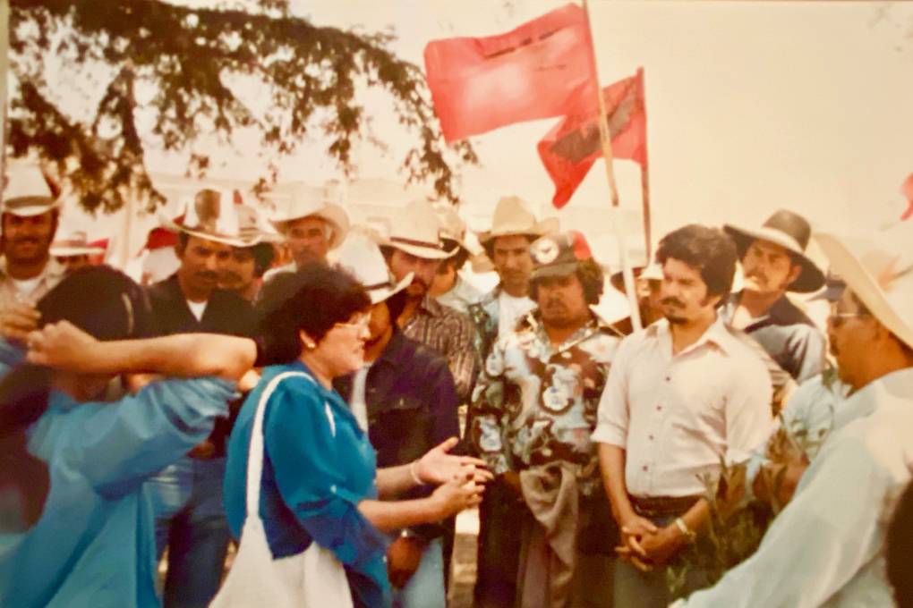 A vintage photo of a woman in a blue dress and glasses talking with a group of men wearing hats and some holding a red flag outside.