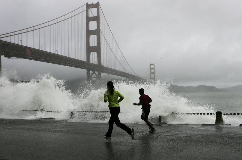 Two people jog along a cement walkway that borders San Francisco Bay on a stormy day. Large waves are crashing over the edge of the walkway. The Golden Gate Bridge is visible in the background.