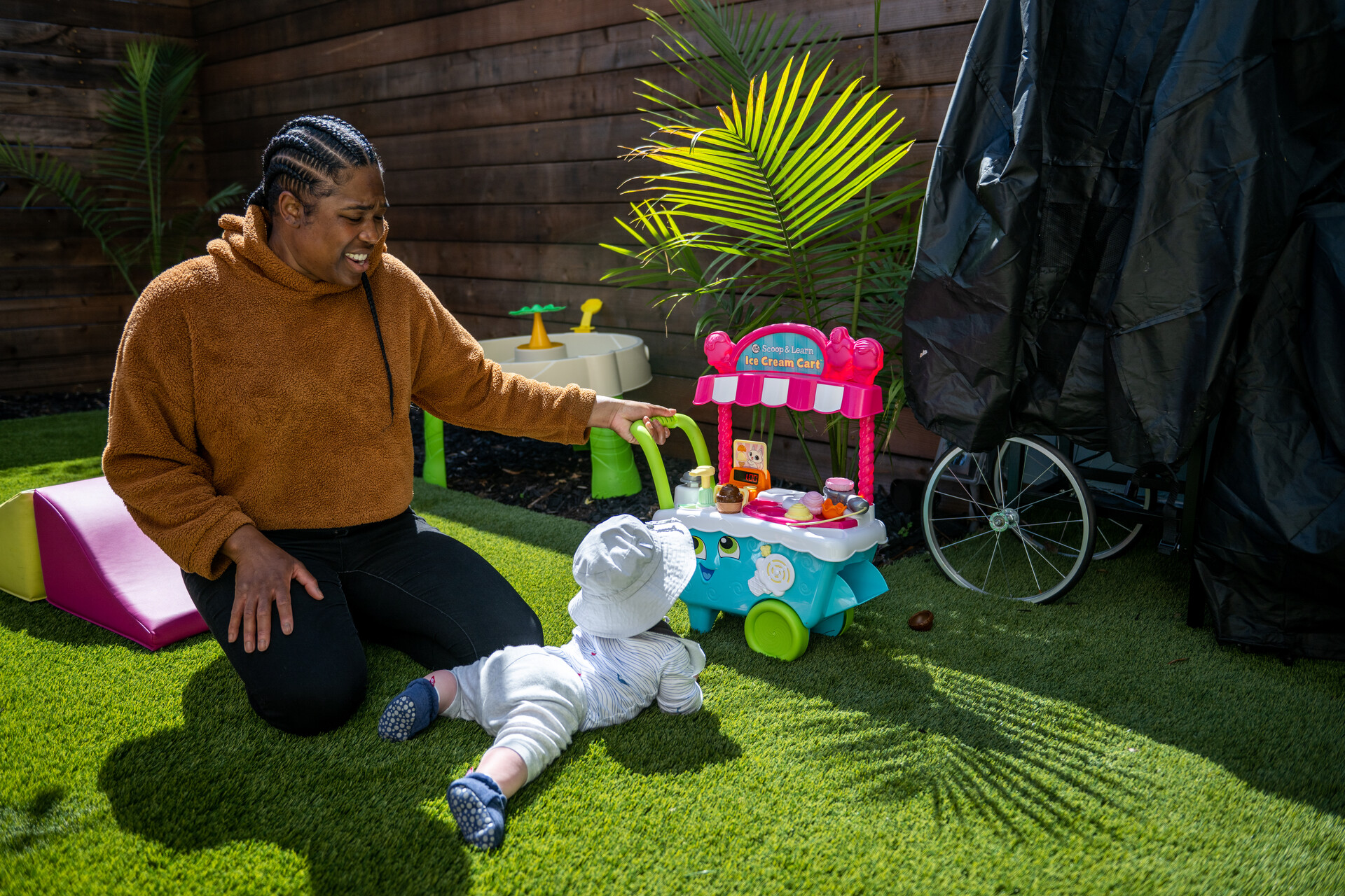 A woman with long, black braids and a brown hoodie plays with a baby in a sunhat in the backyard on a sunny day.