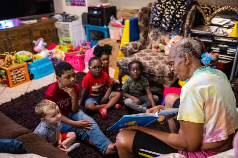 An older woman with gray and brown braids and a rainbow, tie-dye T-shirt reads to a group of children in her living room. Many colorful bins are in the background filled to the brim with toys.