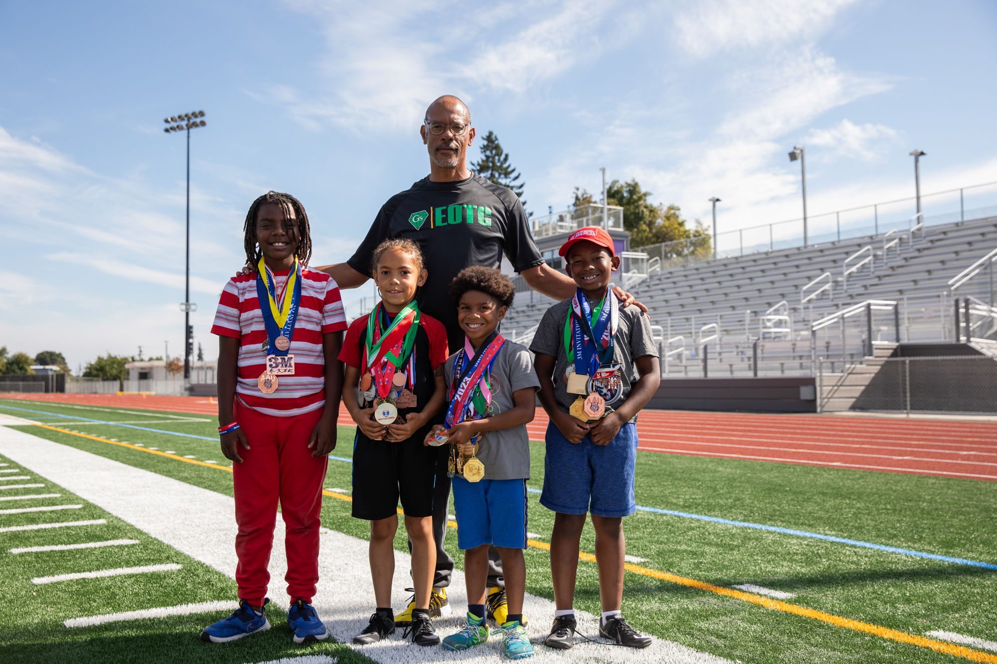 Four children wearing lots of medals pose for a photo with a man on an athletic field.