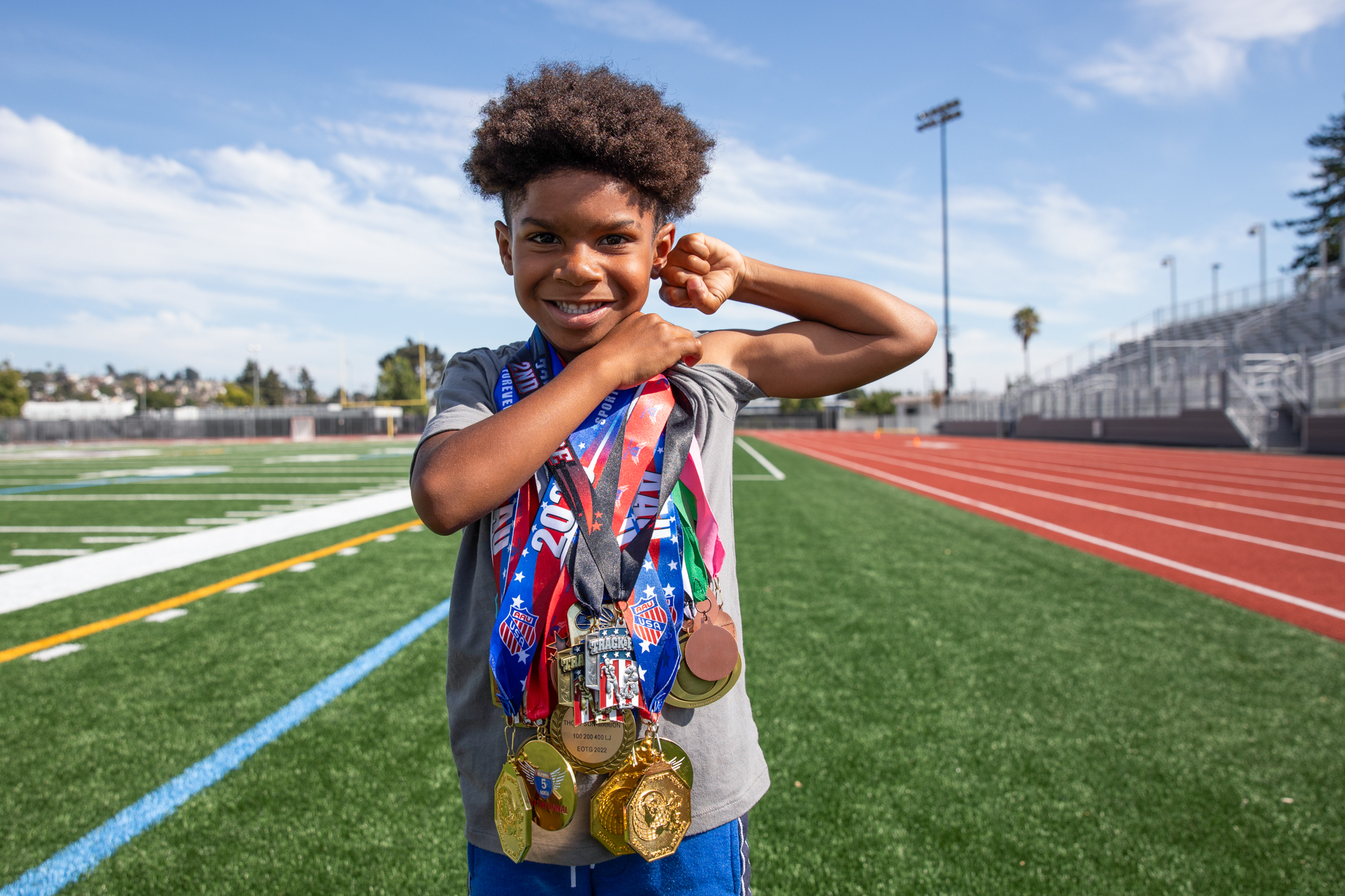 A boy flexes his bicep muscle while posing for a photo with over ten medals hanging from around his neck.