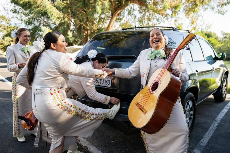 Women wearing ornate white outfits and holding instruments laugh with each other next to a car in a parking lot.