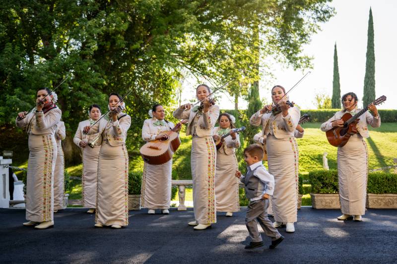 Women wearing ornate white outfits sing and play instruments in an outdoor setting as a child in a suit runs by.