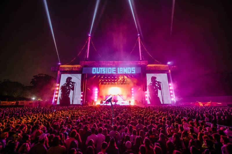 A pink neon-toned nighttime shot of a big crowd standing in front of a stage with a neon sign that says OUTSIDE LANDS.