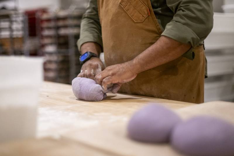 The hands of someone wearing an apron works with purple dough.