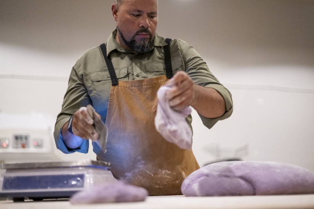 A person with a goatee and wearing an apron works with purple dough.
