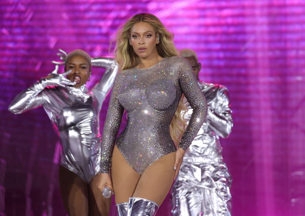 An image of Beyoncé, a Black woman with long honey blonde hair, onstage wearing a silver metallic bodysuit. Dancers are posed behind her, against a purple backdrop.