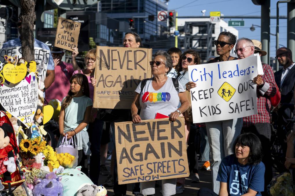 A group of people holds signs that read "Never Again, Safe Streets Now" in an outdoor setting.