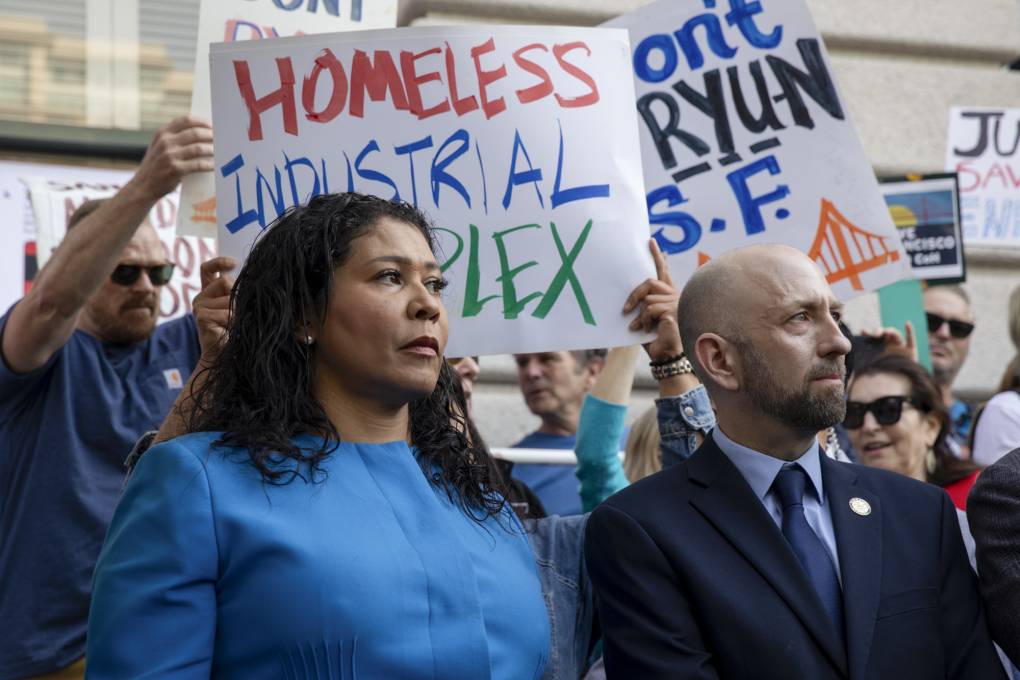 A woman in blue stands next to a man in a suit with signs protesting and supporting homelessness in the background.
