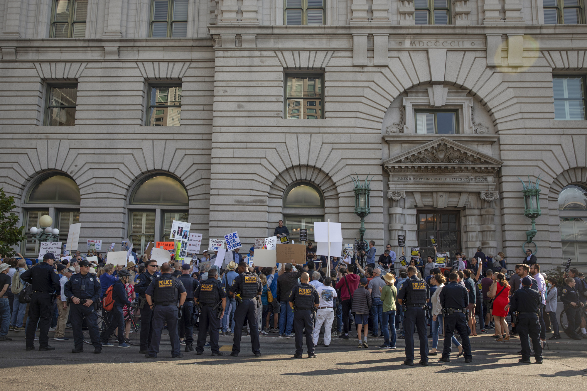 A large crowd with signs gathers in front of a large stone building. A line of police officers stands nearby.